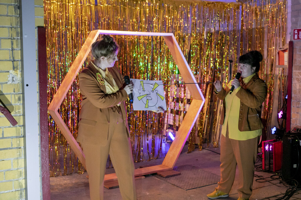 Szenenfoto aus einer Auffuehrung: Zwei personen vor einem goldenen Vorhang auf einer Theaterbuehne.