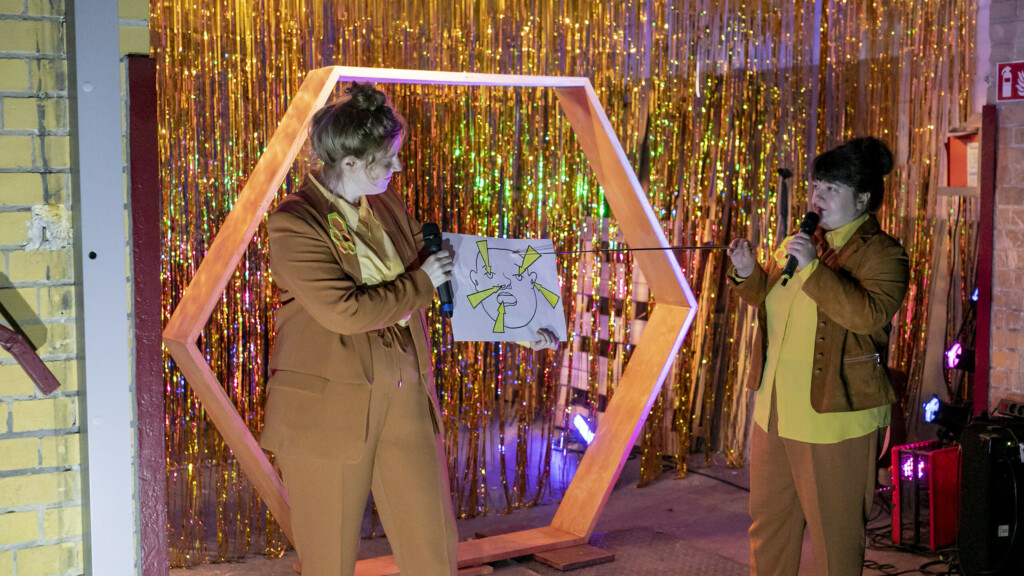 Szenenfoto aus einer Auffuehrung: Zwei Personen vor einem goldenen Vorhang auf einer Theaterbuehne.