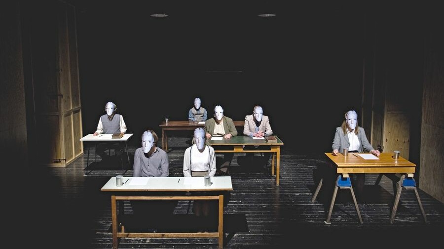 Scene photo of a performance: Seven people are sitting on office tables on a stage wearing masks.