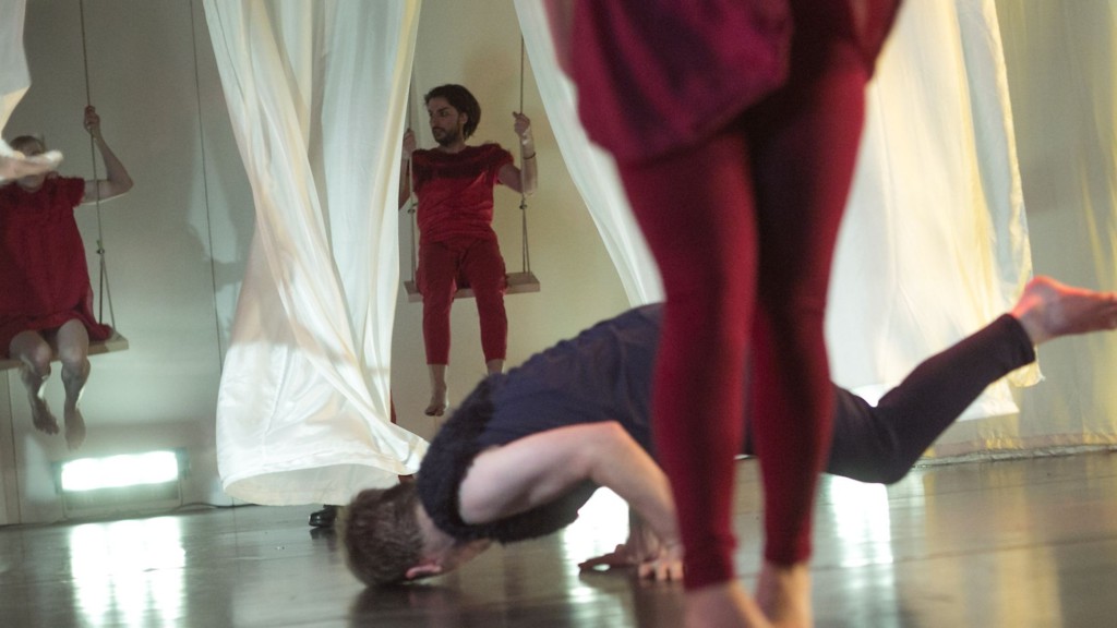 Szenenfoto: Ein Mann dreht sich am Boden, im Vordergrund sind Beine zu sehen, im Hintergrund zwei Menschen auf Schaukeln, die zwischen weissen Vorhaengen schaukeln