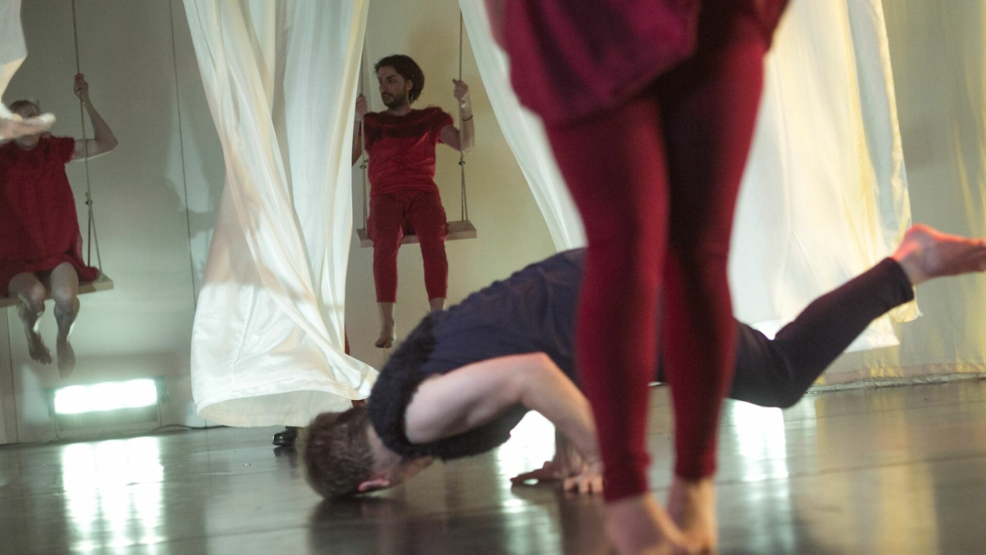 Szenenfoto: Ein Mann dreht sich am Boden, im Vordergrund sind Beine zu sehen, im Hintergrund zwei Menschen auf Schaukeln, die zwischen weißen Vorhaengen schaukeln