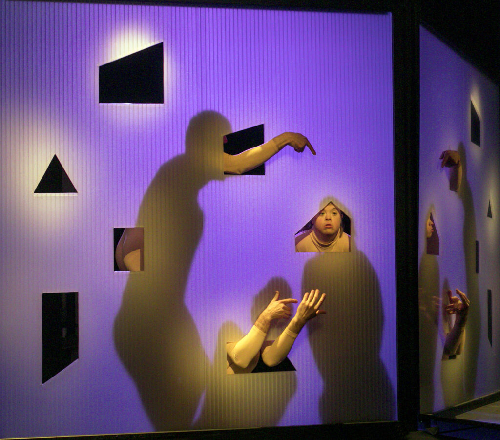 scene photo of a performance: shadows of three people behind a transparent wall on a theatre stage. Some body parts are visible.
