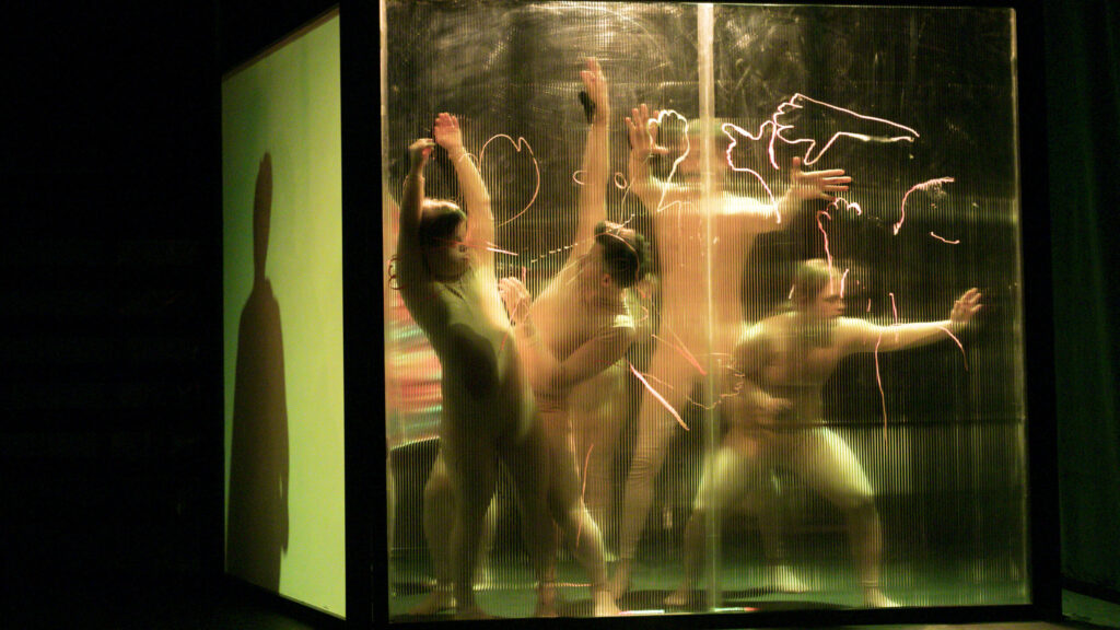 Szenenfoto einer Auffuehrung: Vier Personen hinter einer transparenten Wand auf einer Buehne.