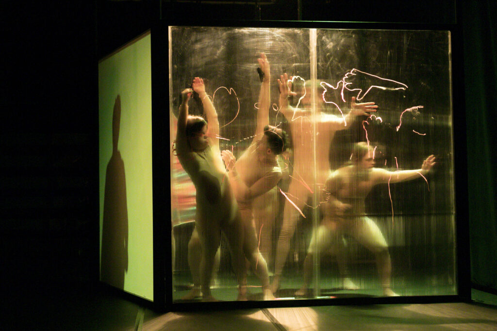 scene photo of a performance: four people behind a transparent wall.