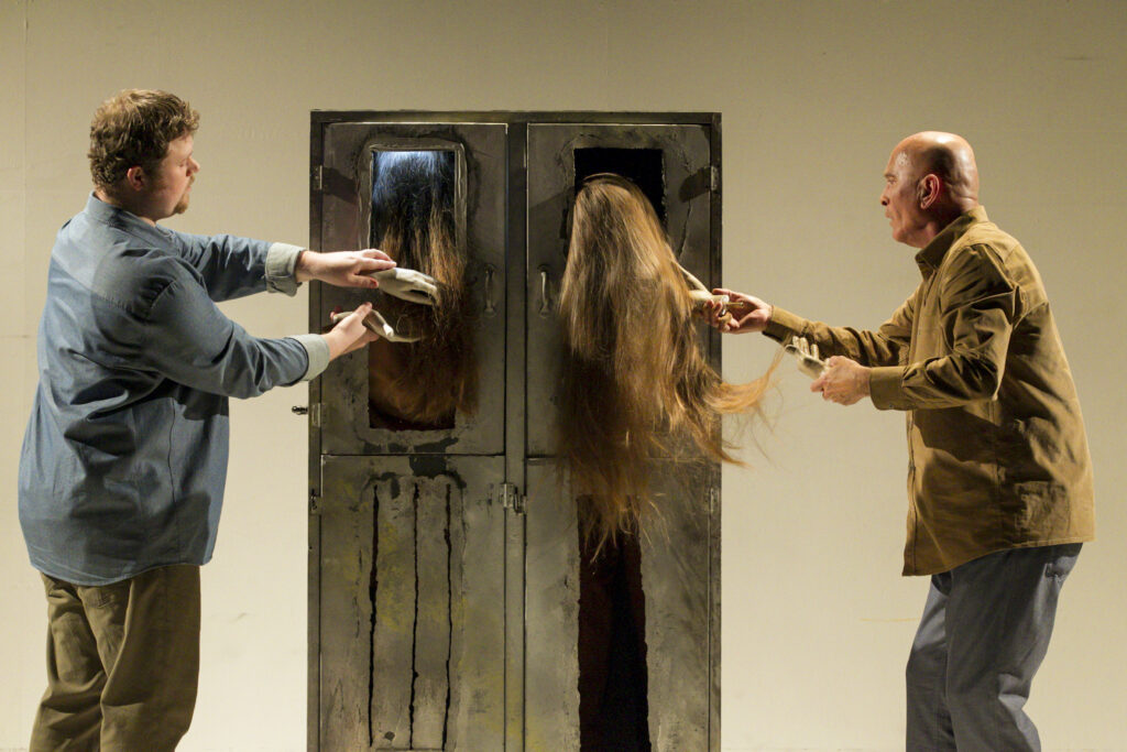 Scene photo of a performance: A metal cupboard on a stage. One men to the left one man to the right with plastic hands. Hair is hanging out of the metal cupboard, which is touched by the hands.