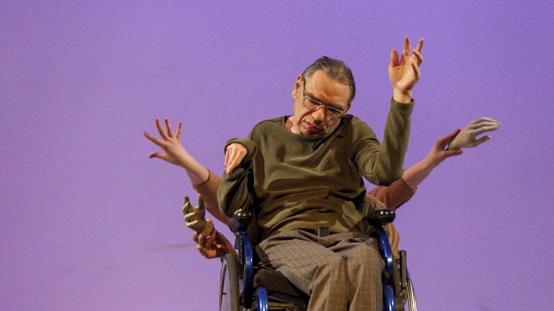 Scene photo of a performance: A man in a wheelchair on a stage. Other people are being him. But you just see their hands.