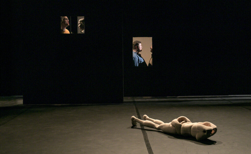 Scene photo of a performance: One puppet without head is lying on a stage. Behind you can see people in little windows.