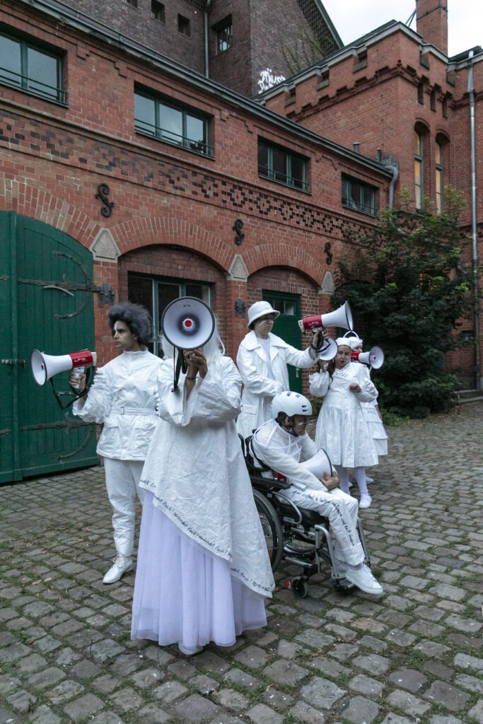 Szenenfoto einer Auffuehrung: Mehrere Personena uf einer Buehne in weissen Kostümen mit Megafonen.