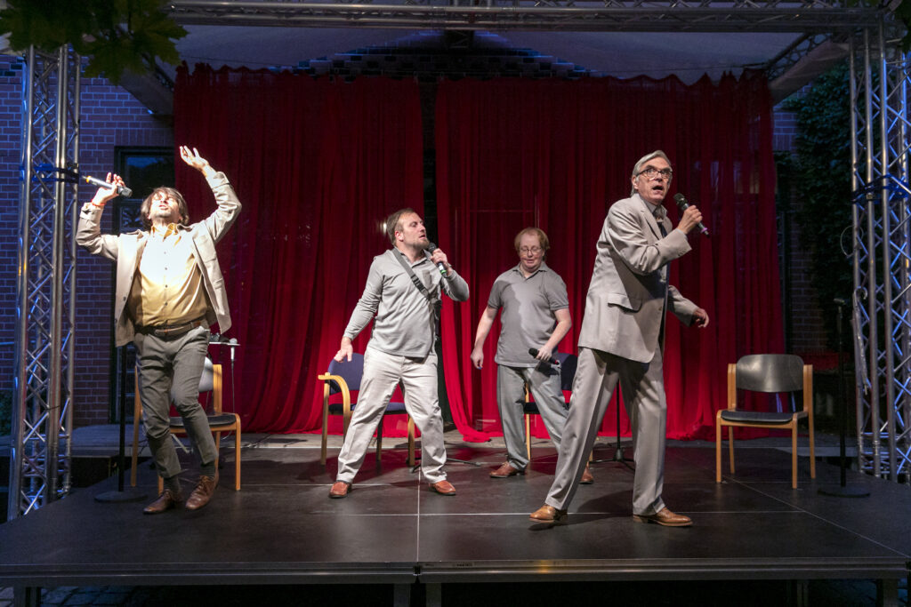 Scene photo of a performance: Four men in suits on a stage.