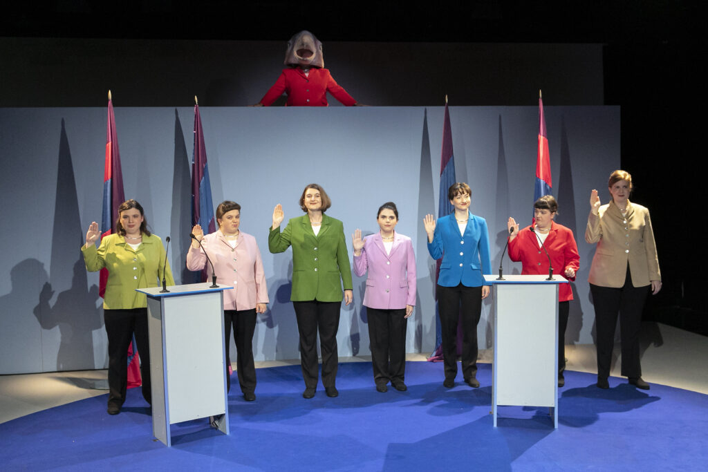 Scene photo of a performance: seven performers wearing blazers and holding up there right hand. In the middle a person with eagle mask.