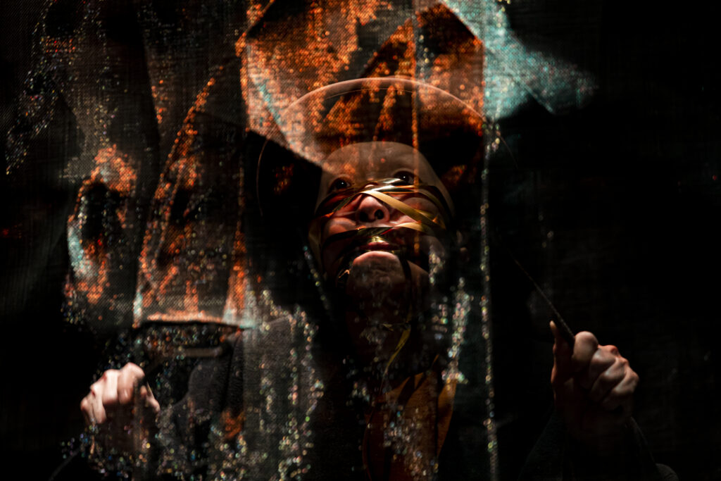 Scene photo of a performance: woman with a hat behind a seethrough metal curtain