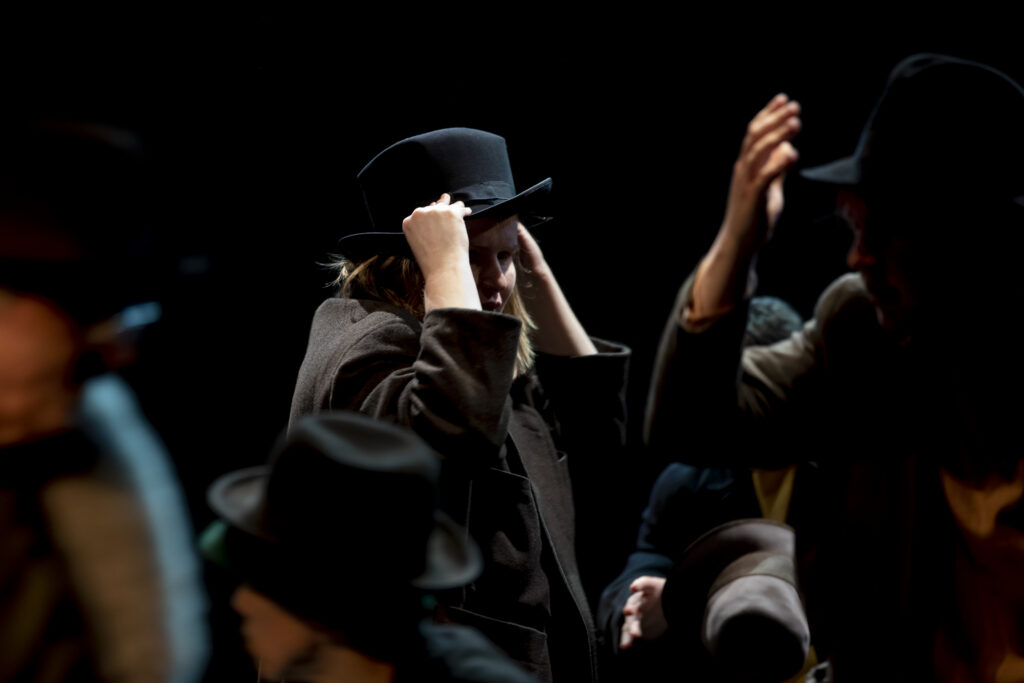 Szenenfoto einer Auffuehrung: Eine Person auf einer Bühne hält ihren Hut fest. Vorn im Bild rechts sind Haende anderer Personen zu sehen.