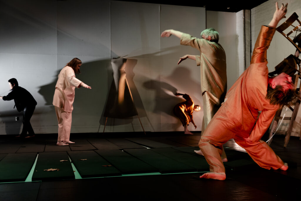 Scene photo of a performance: Some people on a stage. They dance. In the middle a statue of felt.
