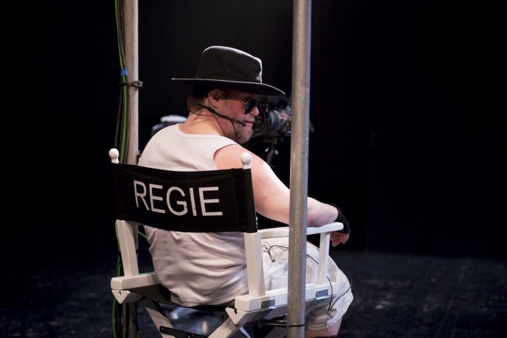 Scene photo of a performance: One man is sitting in a directors chair.