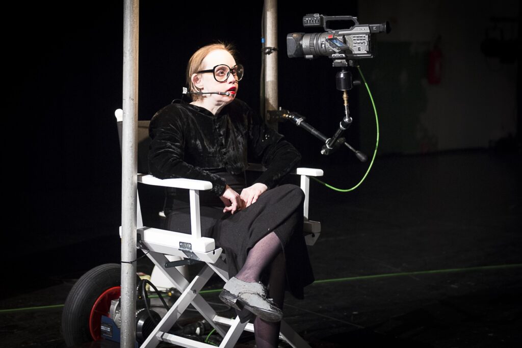 Scene photo of a performance: One woman is sitting in a directors chair.