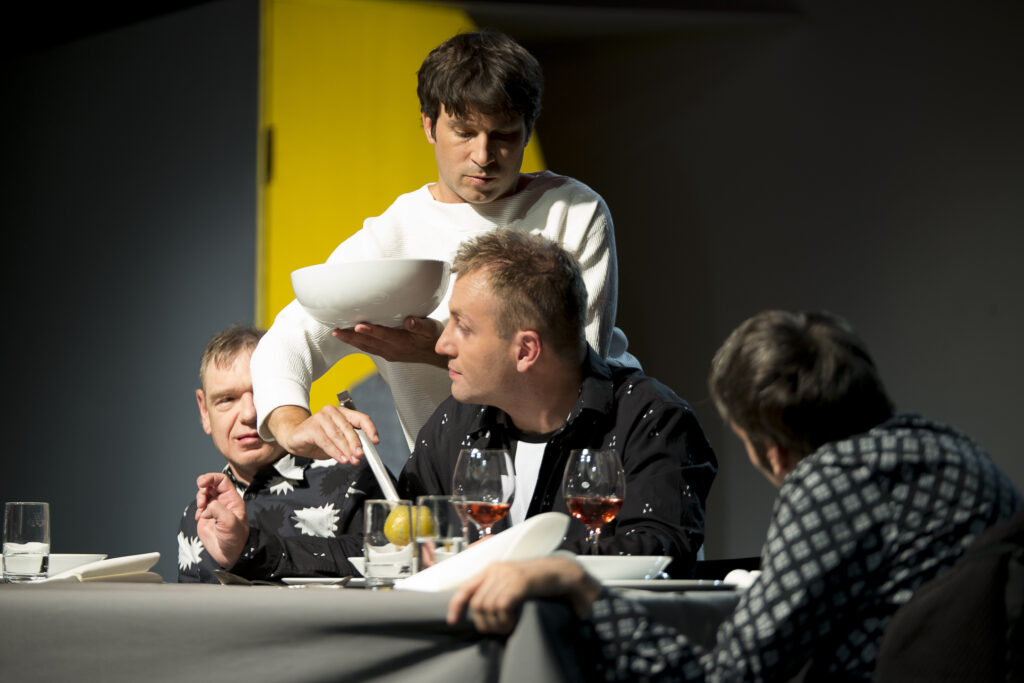 Szenenfoto einer Auffuehrung: drei Personen sitzen am Tisch ein einer Buehne. Einer steht zwischen den beiden ersten Personen von links und schenkt ein Getraenk ein.