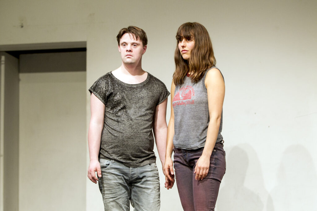 Scene photo of a performance: a man and a woman next to eachother on the stage. They are looking in the direction of the audience.