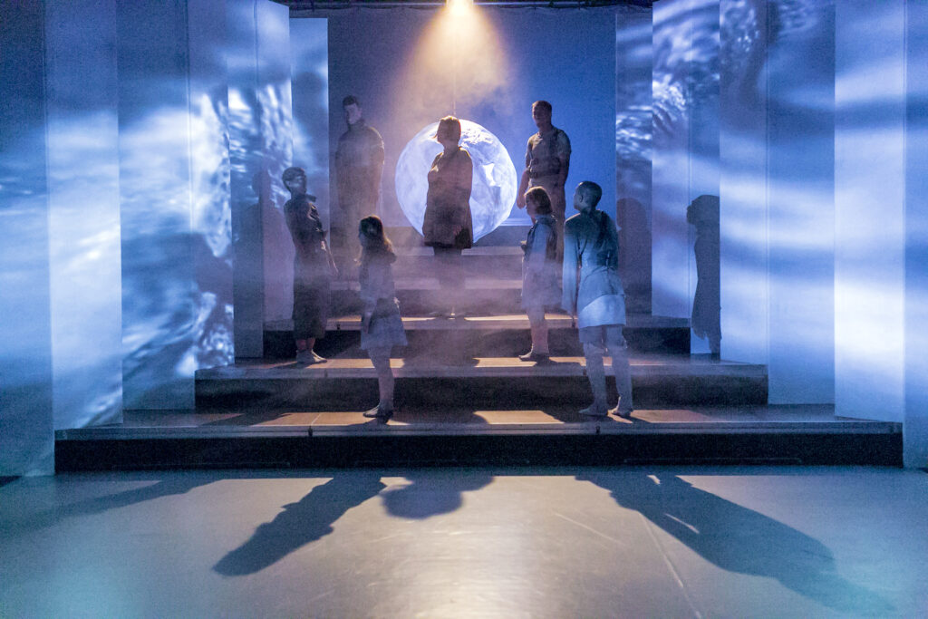 Szenenbild einer Auffuehrung: Mehrere Personen auf iener Bühnen stehen versetzt auf einer Treppe.