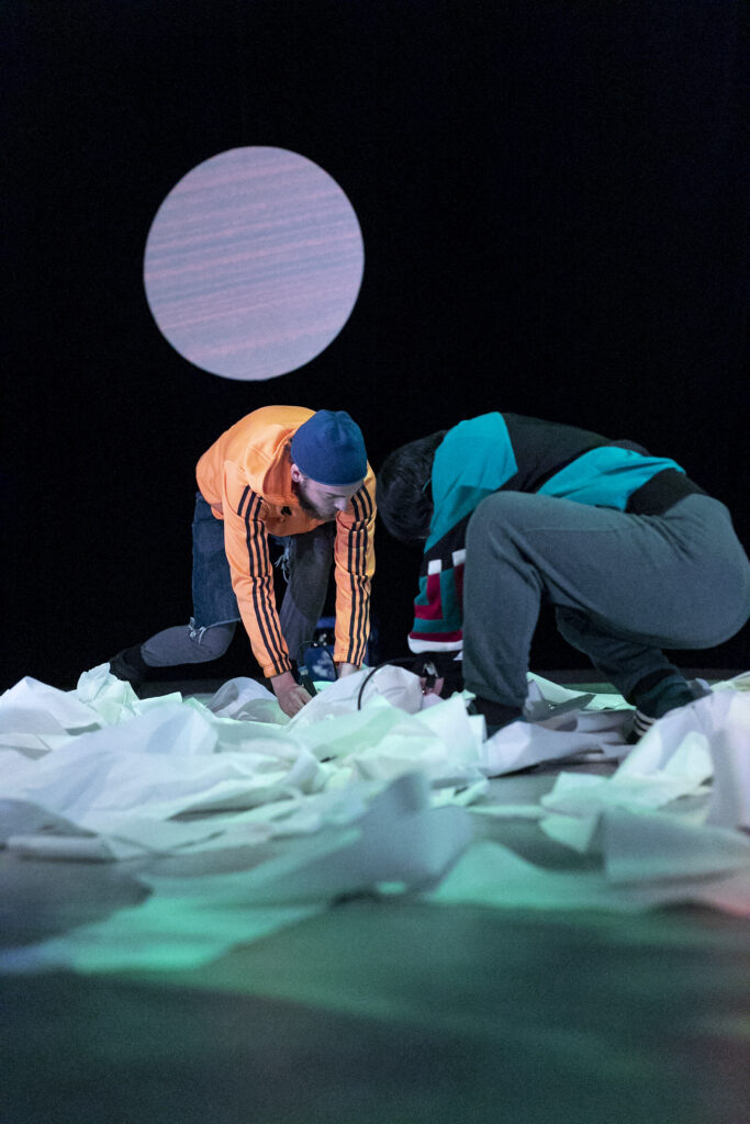 Szenenfoto einer Auffuehrung: Zwei Personen gegenueber gebeugt auf einer Theaterbuehne. Um sie herum Kuchenpapier mit dem sie interagieren.