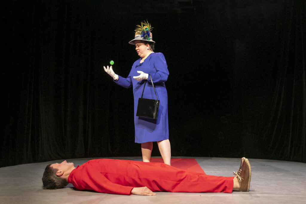 Szenenfoto einer Auffuehrung: Eine Person liegt auf dem Rücken auf dem Boden in einem roten Gewand, dass sich bis zum Buehnenende wie ein roter Teppich erstreckt. Sie schaut zu der Person hinter ihr. Diese wird einen Flummi mit der rechten Hand in die Luft.