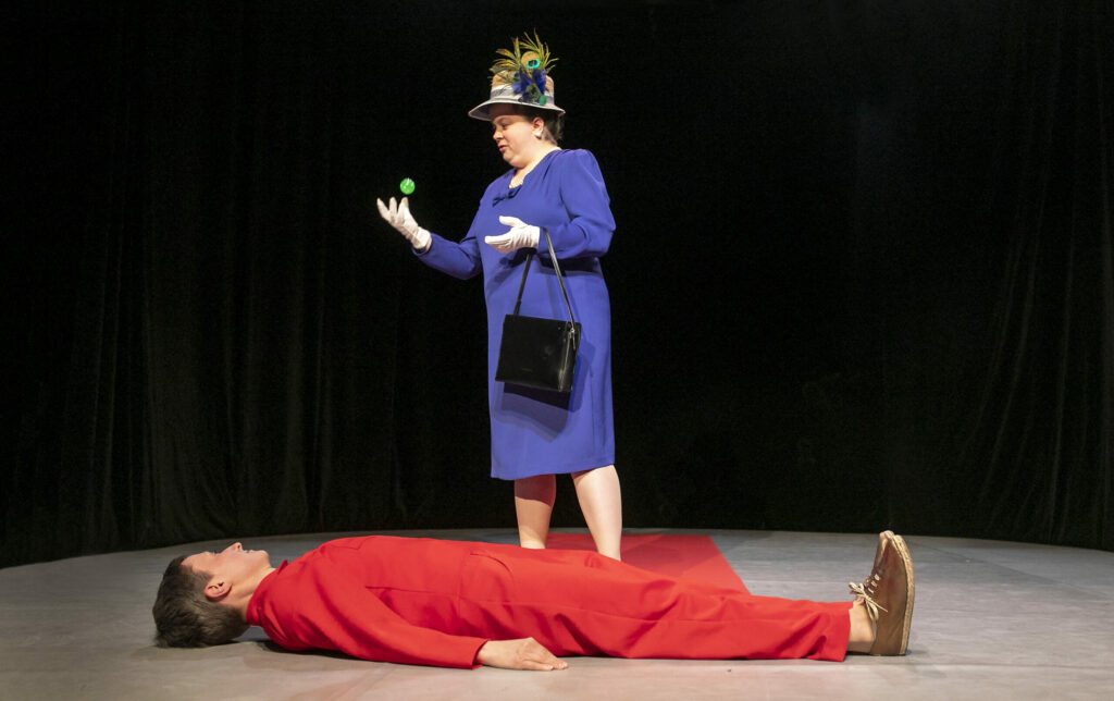 Scene photo of a performance: One person is lying on her back on the ground. She is looking at the other person behind her who throws a bouncy ball in the air with her right hand.