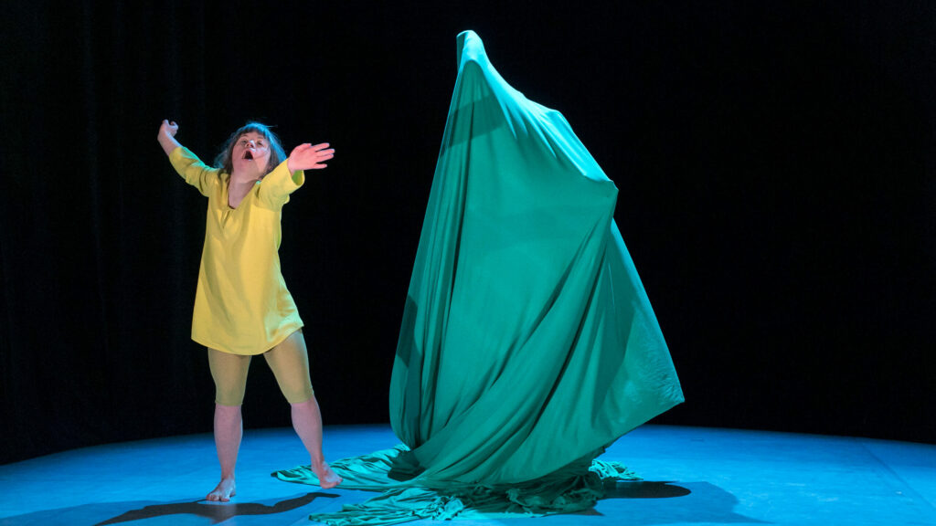 Scene photo of a performance: One person with wide open arm on the left. On the right side a person under green fabric.