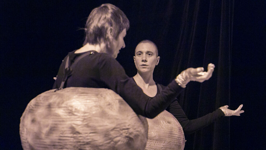 Scene photo of a performance: Two people on a stage. Both have their arms tilted. They wear a ball as a costume around their body.