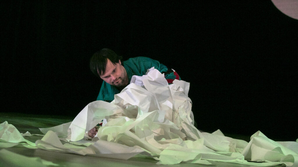 Scene photo of a performance: One person is crawling on a theatre stage. A pile kitchen towels in front of her.