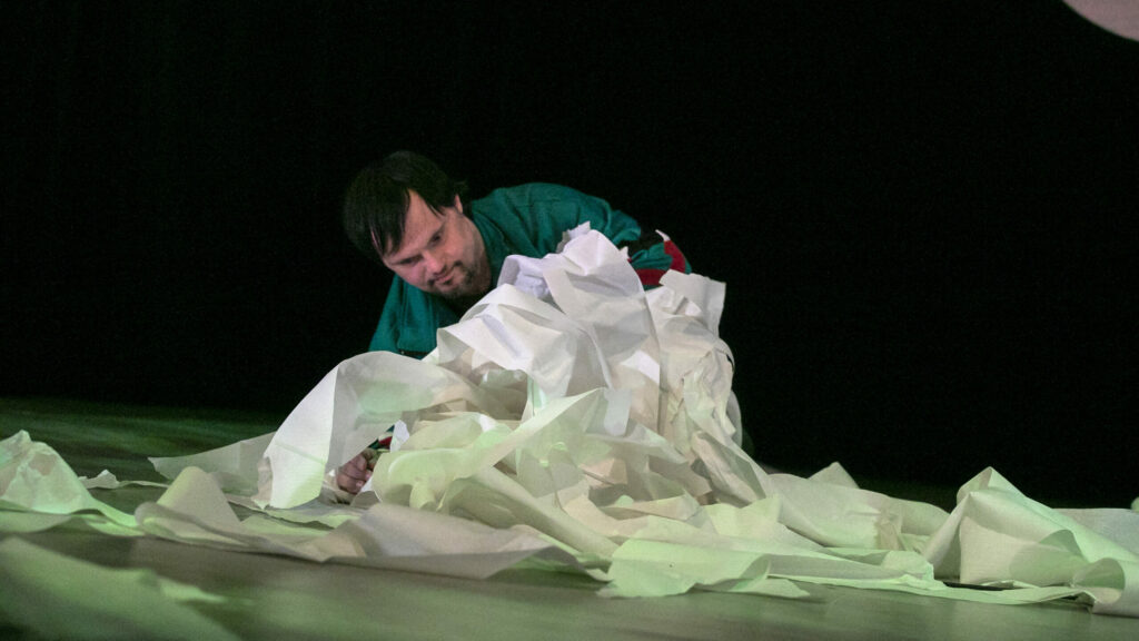 Szenenfoto einer Auffuehrung: Eine Person kriechend auf einer Theaterbuehne. Ein Haufen abgerolltes Kuechenpapier vor ihr.