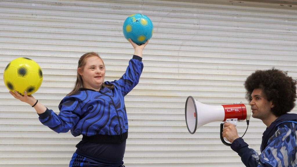 A person is holding a megafon. The other person is holding her hands in the air and is holding a ball in every hand.