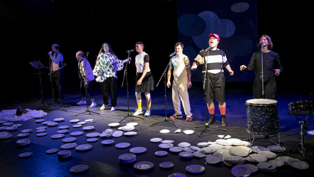 Scene photo of a performance: Seven People next to each other. Microphones in front of them.