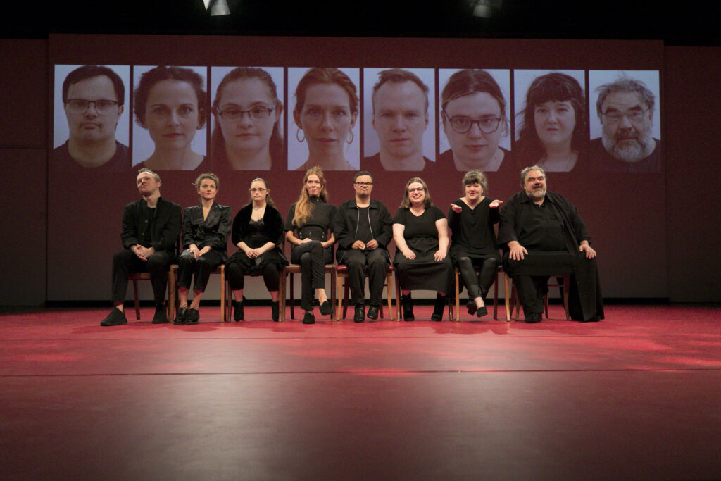 Szenenfoto einer Auffuehrung: Mehrere Personen sitzen in einer Reihe nebeneinander auf einer Theaterbühne. Im Hintergrund eine Projektion mit verschiedenen Gesichtern.