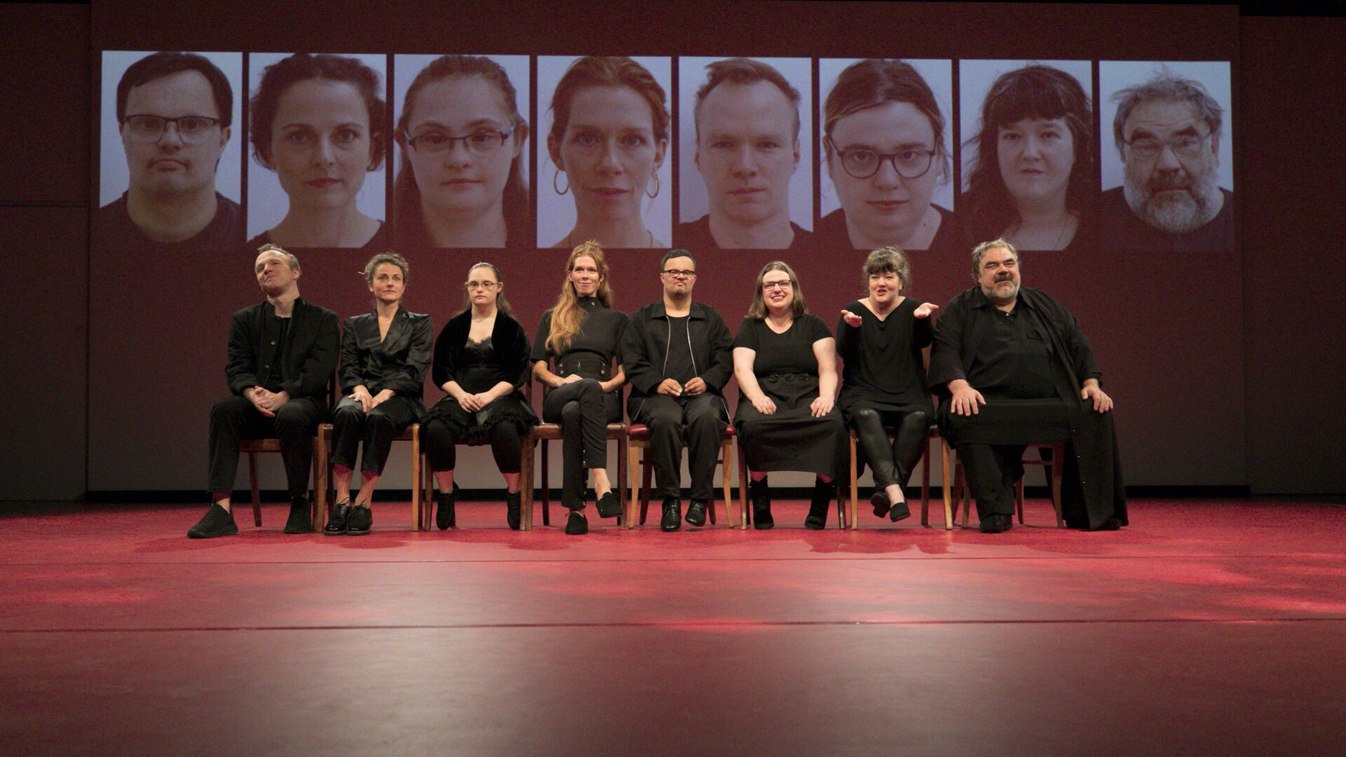 Szenenfoto einer Auffuehrung: Mehrere Personen sitzen in einer Reihe nebeneinander auf einer Theaterbühne. Im Hintergrund eine Projektion mit verschiedenen Gesichtern.