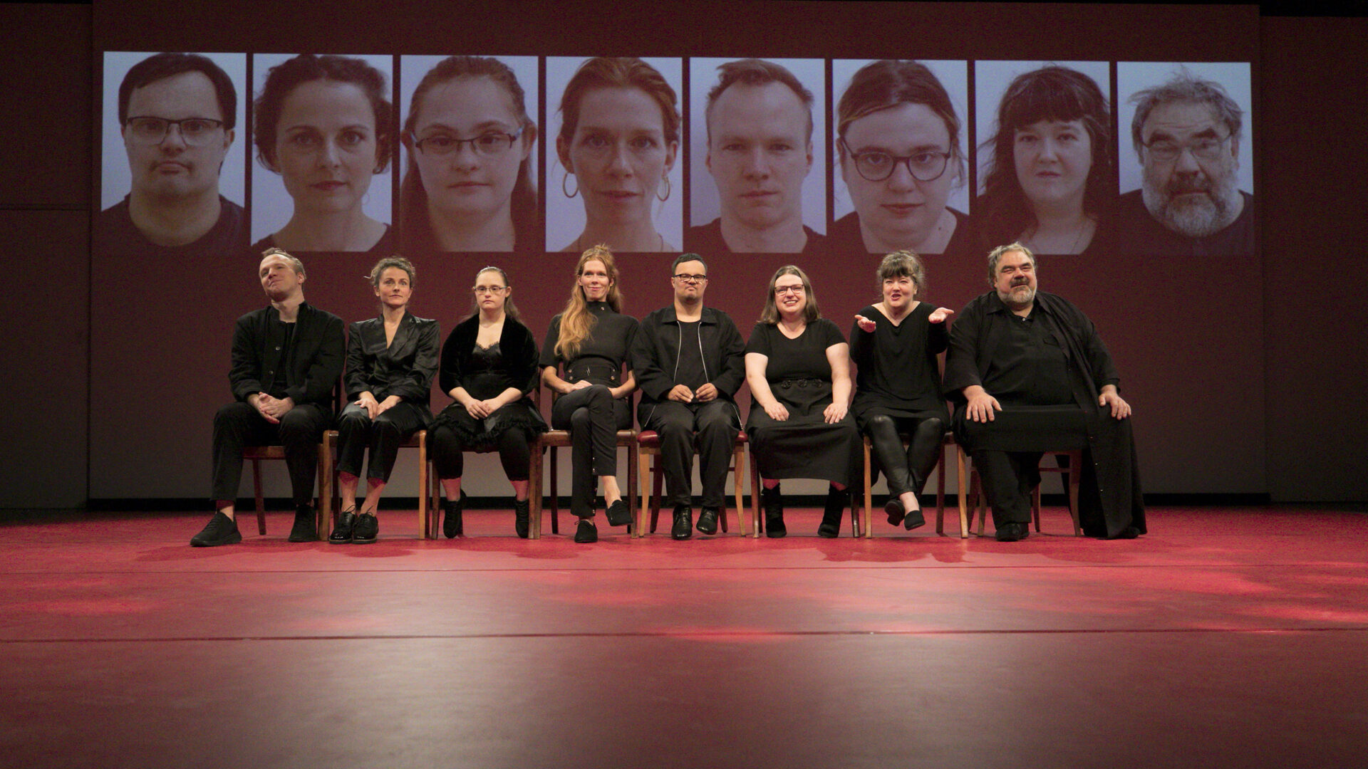 Scene photo of a performance: A couple people are sitting in a row next to each other. In the background a projection with different faces.