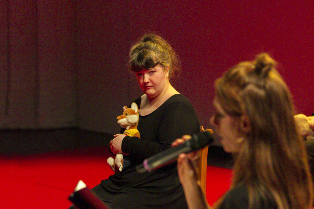 Scene photo of a performance: A person sits on a chair. She has a plush fox. In the foreground another person, who is talking using a microphone.