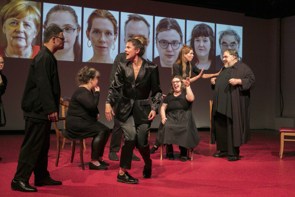 Scene photo of a performance: A couple people on a stage. Two are sitting. The others are standing. The interact with each other.