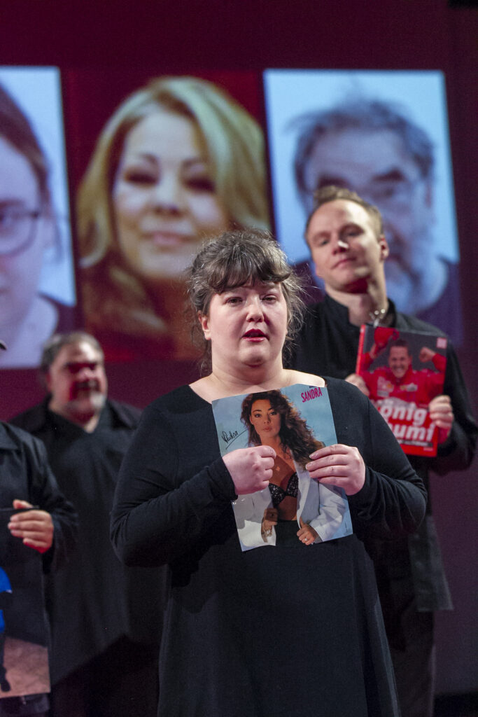 Scene photo of a performance: A person is holding a piece of a newspaper with their idol. In the background zwo other people who do the same.