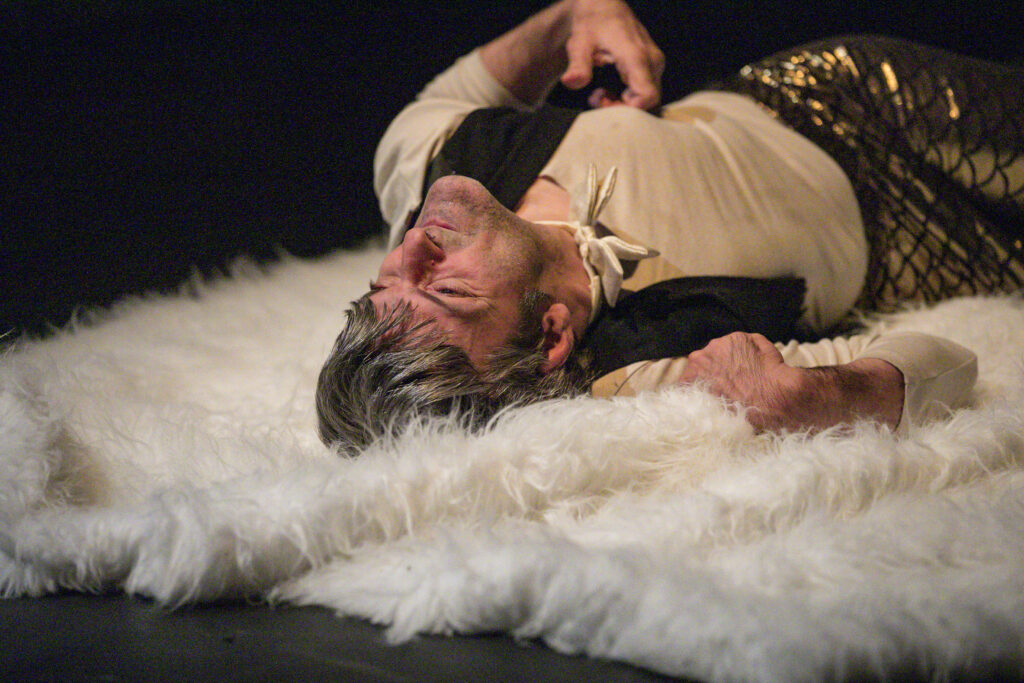 Scene photo of a performance: One man lies on his back on a fur carpet.