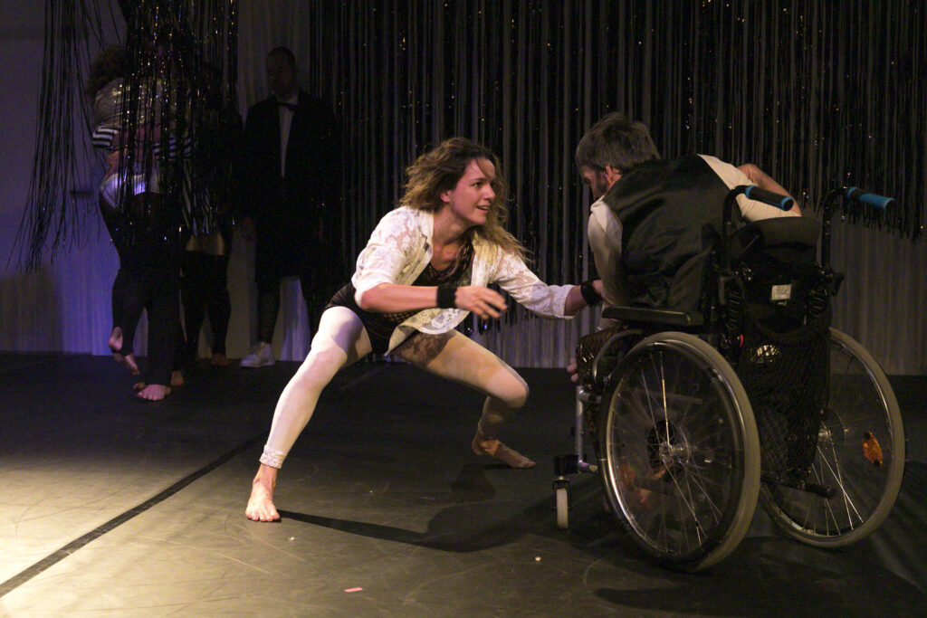 Szenenfoto einer Auffuehrung: Eine Taenzerin links. Rechts ein Mann im Rollstuhl.
