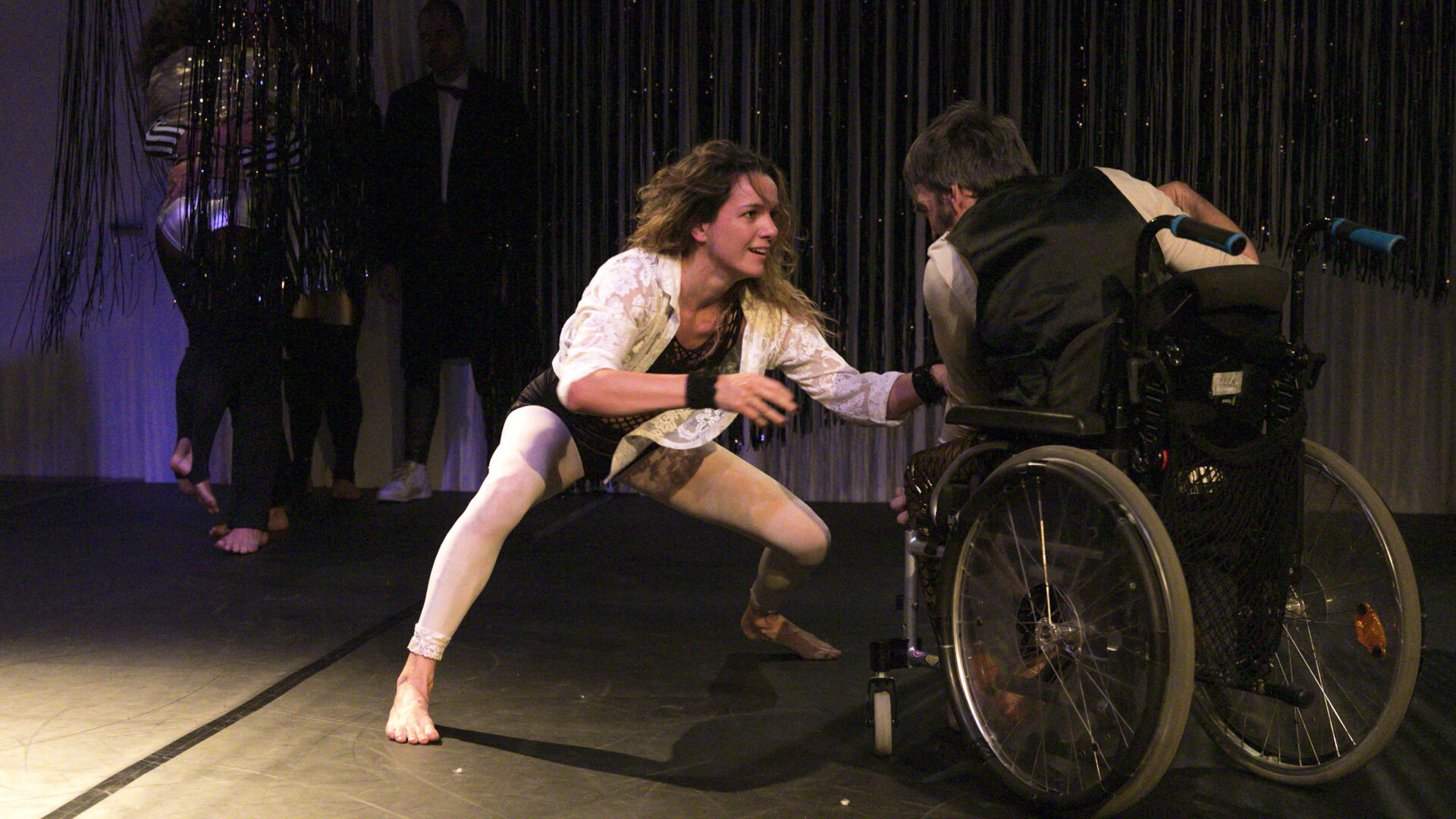 Scene photo of a performance: One dancer on the left. One the right side a man in a wheelchair.