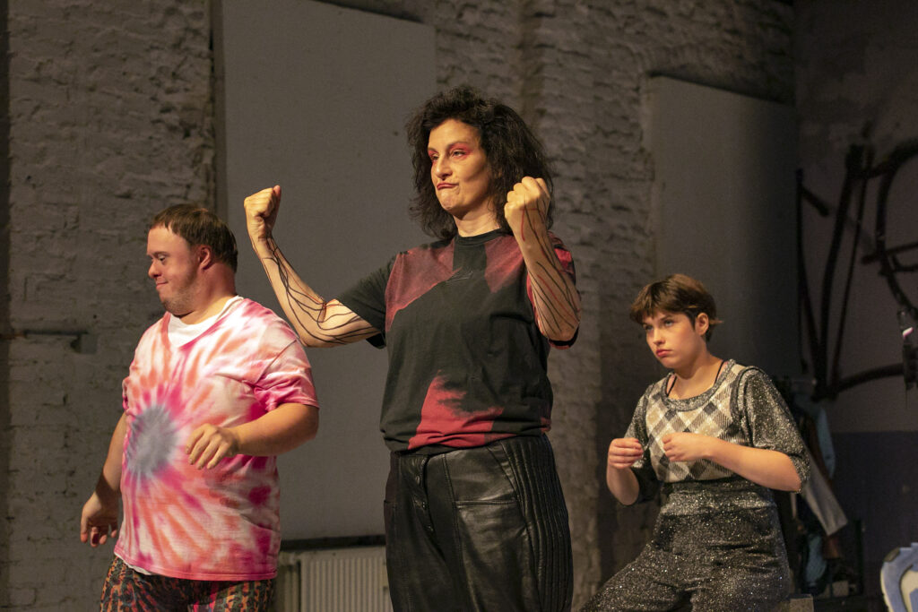 Scene photo of a performance: Three people on a stage. One in the middle with tilted arms.