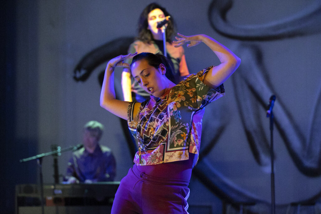 Scene photo of a performance: one Person is dancing with arms up on the foreground. One is in the background talking into a microphone.