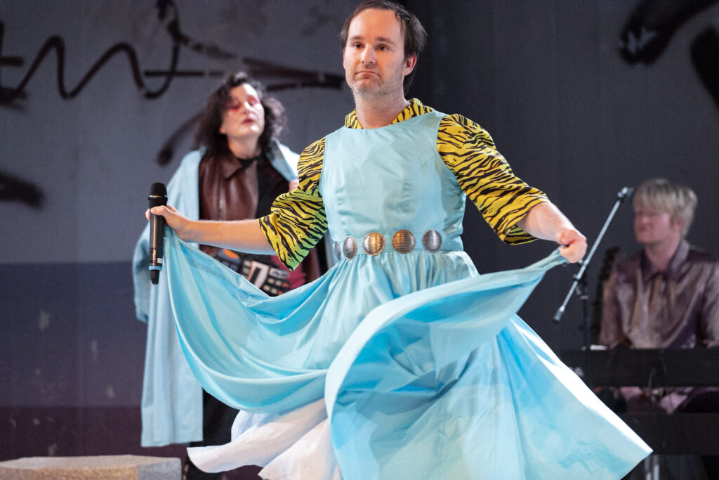 Scene photo of a performance: One man with a bright blue dress on a stage