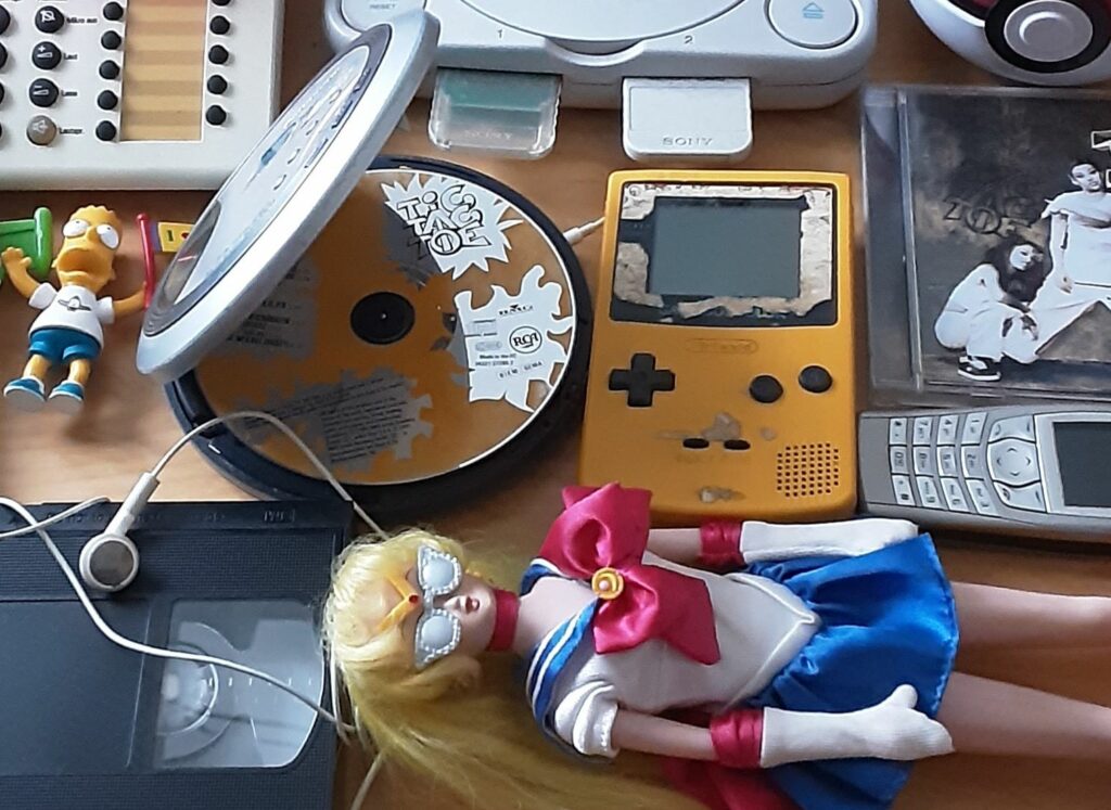 Eine Sammlung von Dingen aus den 1990ern: Discman, Gameboy, Nokia-Handys, ein Tastentelefon, eine Sailor-Moon-Puppe, CDs.