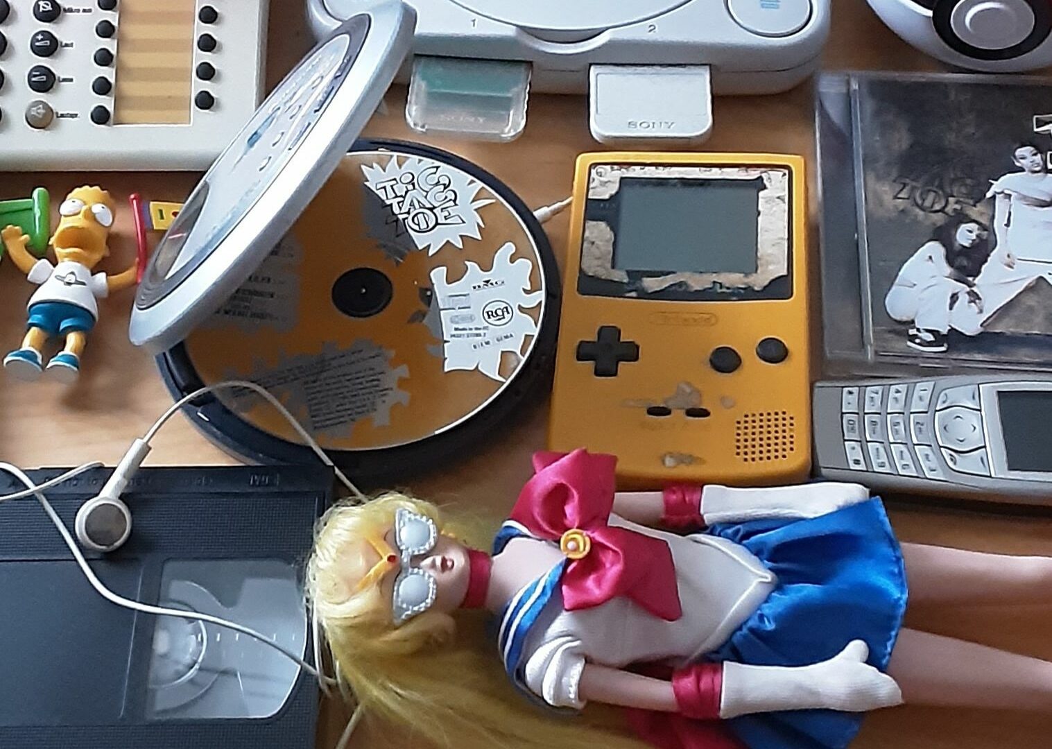 Eine Sammlung von Dingen aus den 1990ern: Discman, Gameboy, Nokia-Handys, ein Tastentelefon, eine Sailor-Moon-Puppe, CDs.