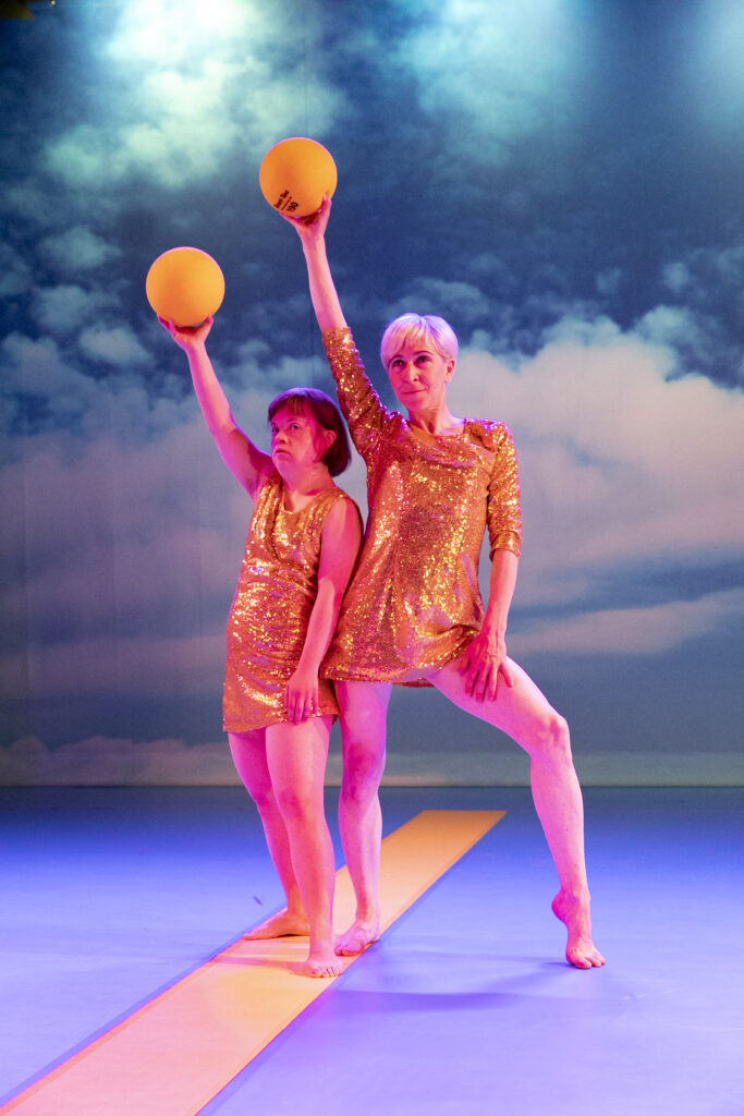 Two people are standing in front of an artificial cloud background. They are illuminated reddish and wear golden costumes. Both have a yellow ball in their hands.