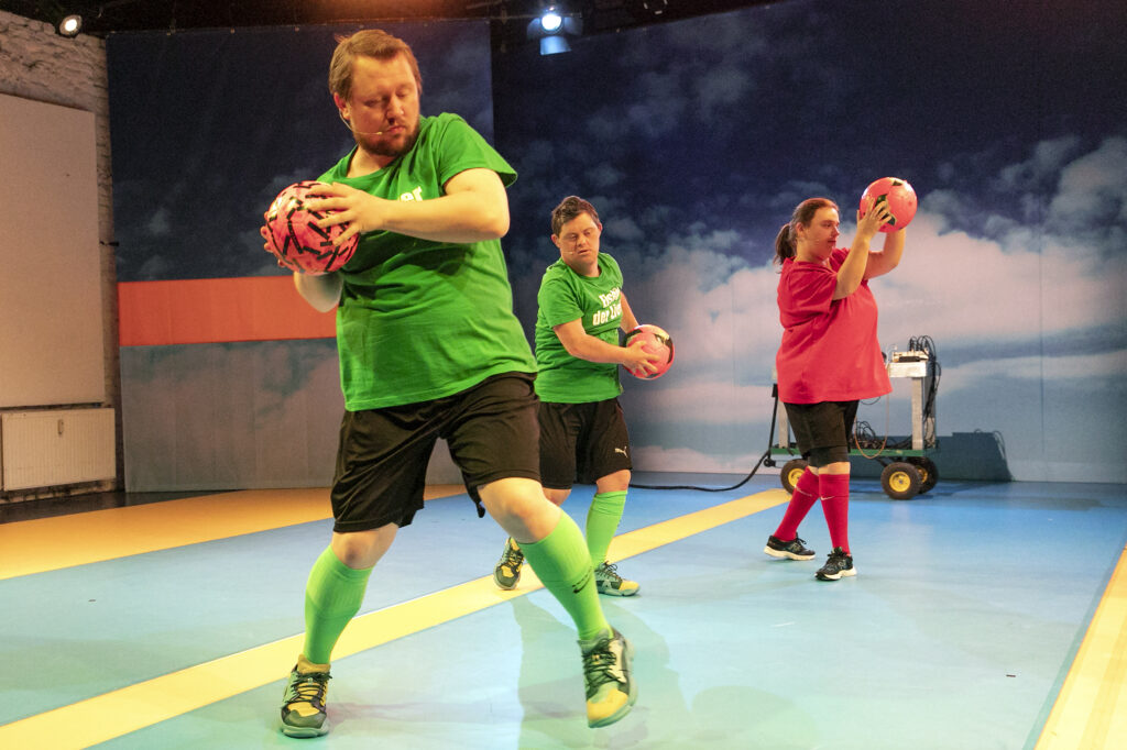 Three people stand on a stage holding pink balls in both hands.