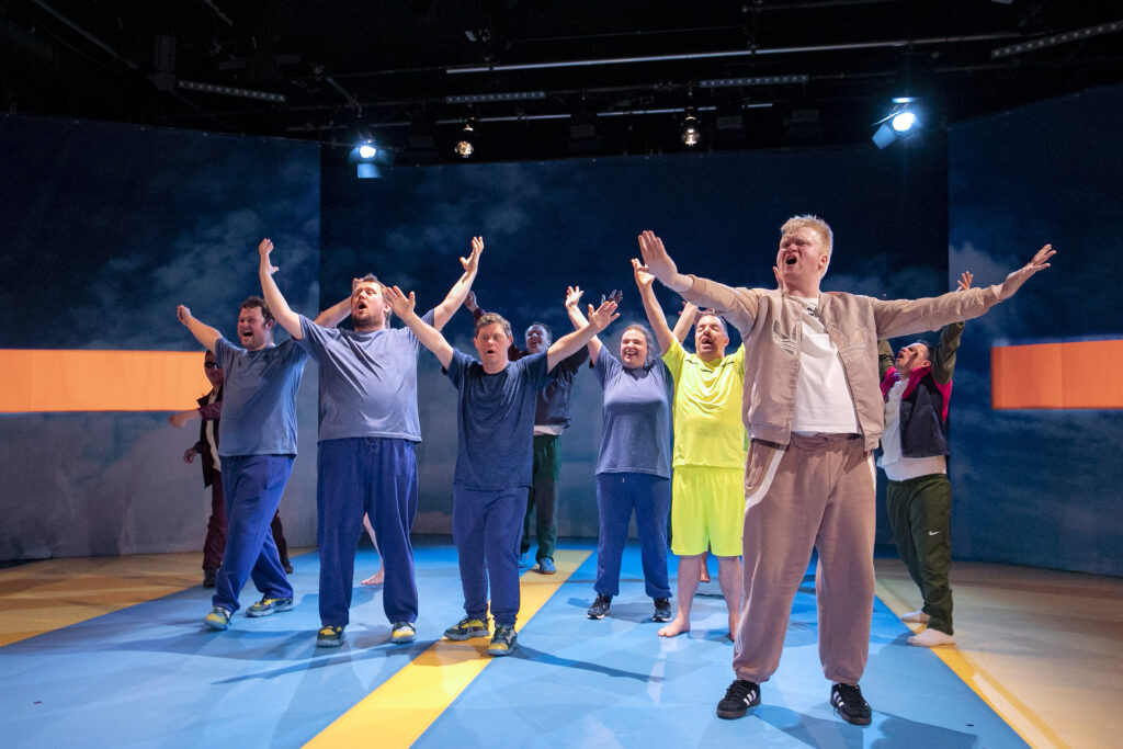 A large group of people stands on a stage on a blue floor with a wide yellow stripe. They seem to be shouting and stretching their arms from themselves.