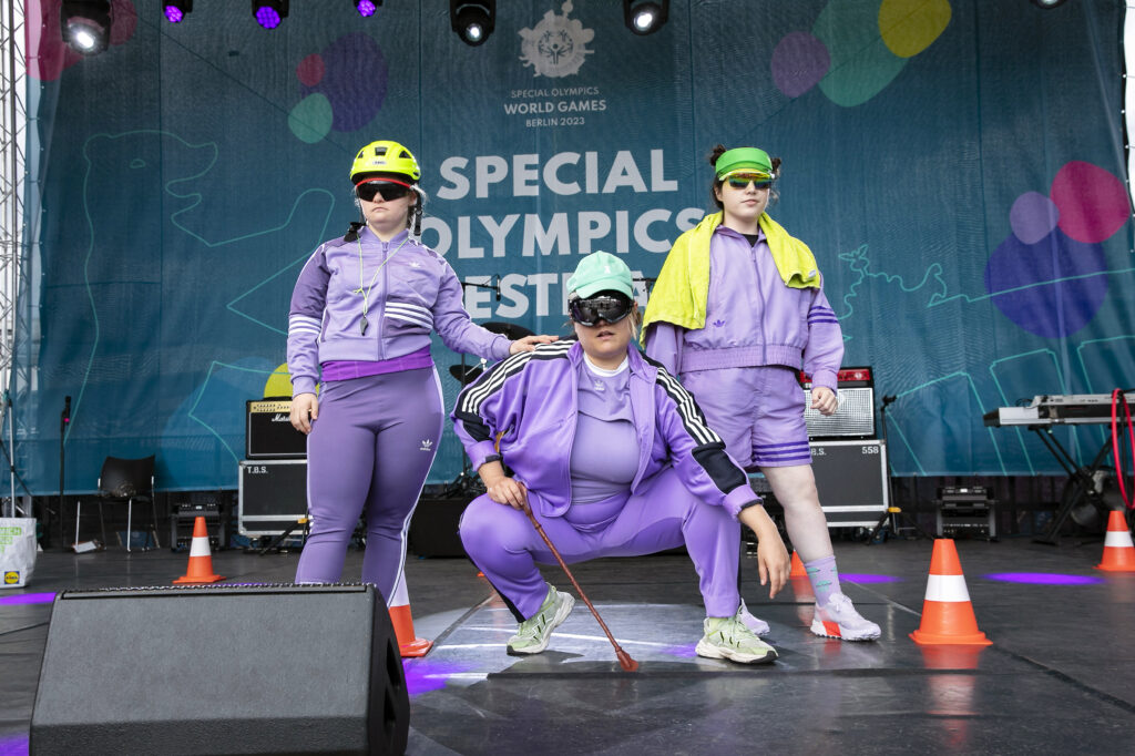 Three people stand on a stage. The person in the middle kneels, the others stand to his left and right. All are wearing lilac sports suits and sunglasses.