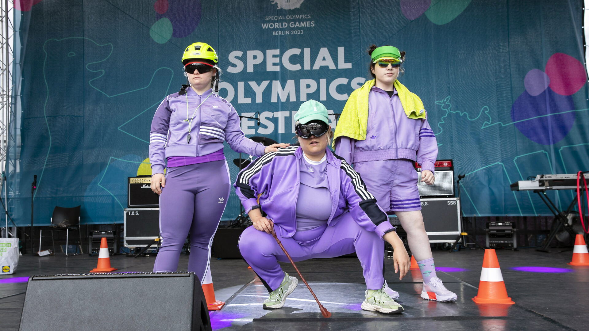 Three people stand on a stage. The person in the middle kneels, the others stand to his left and right. All are wearing lilac sports suits and sunglasses.